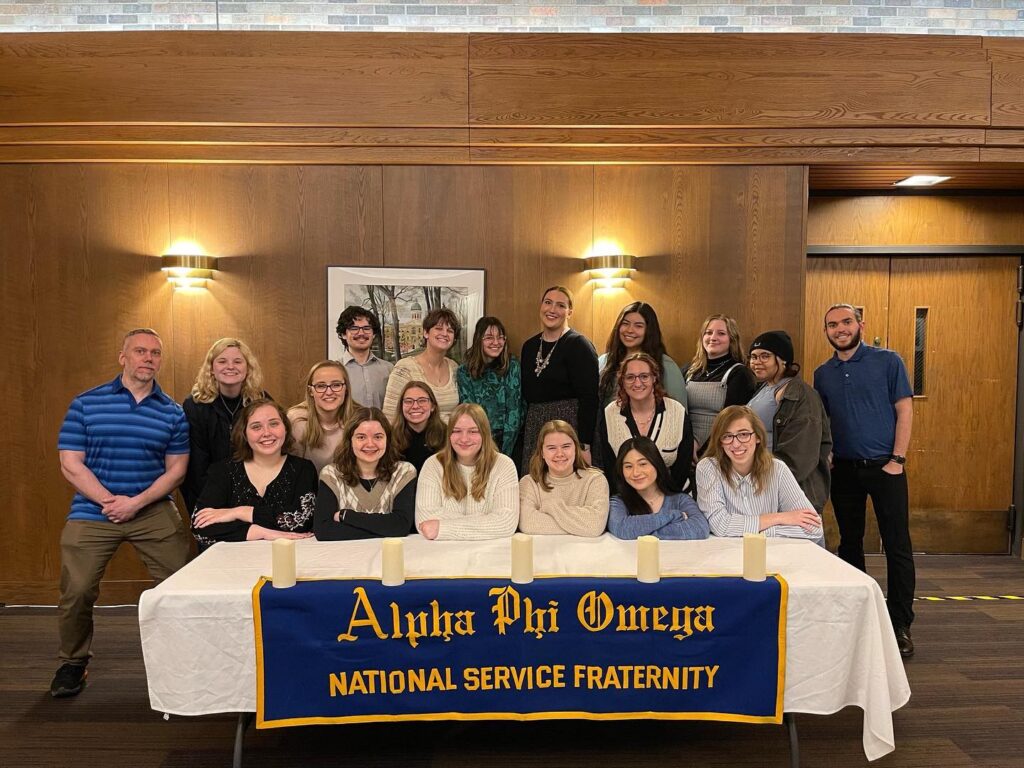 Lambda Rho chapter of Alpha Phi Omega at Augustana College poses as a group behind a table with a blue and gold banner and candles after their Spring 2023 New Member Ceremony.